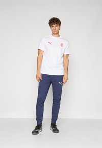 Hvid t-shirt med rødt logo på brystet, parret med marineblå joggers med hvidt logo på venstre ben. Sorte sportssko.