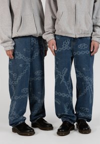 Loose blue denim jeans feature a pattern of chains, brass knuckles, and safety pins in gray. Paired with black shoes and a gray hoodie.