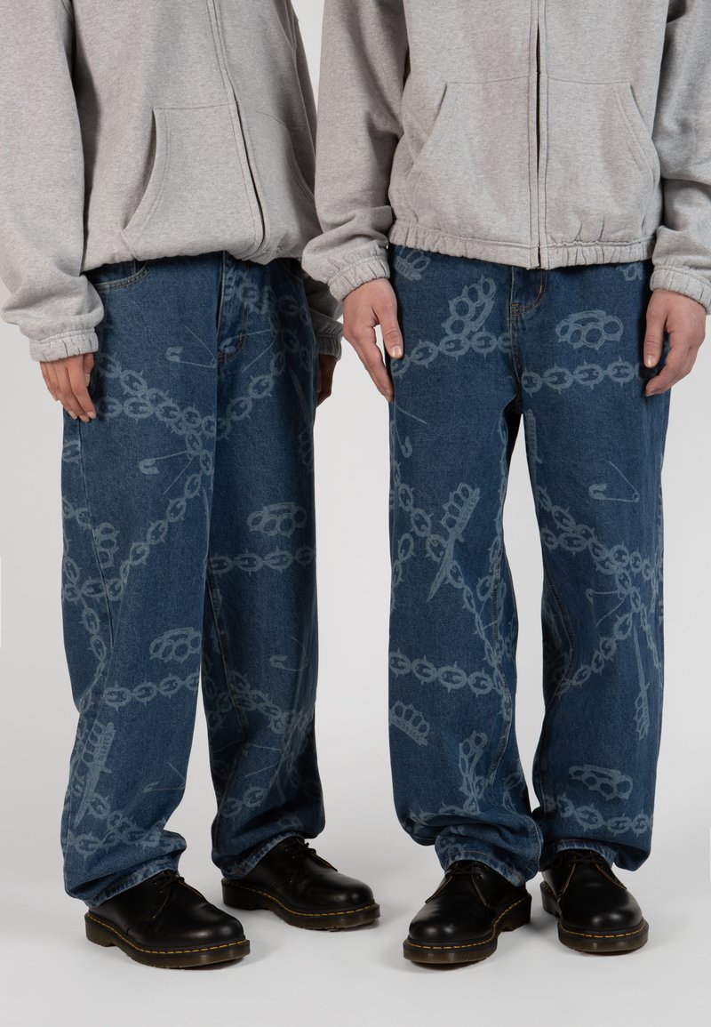 Loose blue denim jeans feature a pattern of chains, brass knuckles, and safety pins in gray. Paired with black shoes and a gray hoodie.