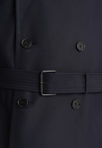 Granatowy płaszcz trencz z dwurzędowym zapięciem, dużymi czarnymi guzikami oraz teksturowanym paskiem z metalową prostokątną klamrą.