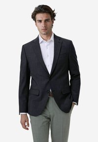 Dunkelkarierter Blazer aus glattem Stoff, mit zwei vorderen Taschen, mit geschlitzten Revers und einem Ein-Knopf-Verschluss, kombiniert mit hellen Hosen.