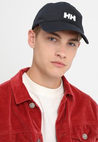 Marineblauwe baseballpet met wit geborduurd logo, gebogen rand en stoffen textuur. Model draagt een rode corduroy jas over een witte shirt.
