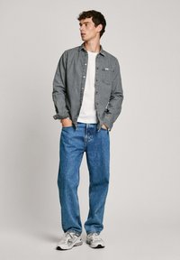 Grijze button-up shirt over een wit T-shirt, gecombineerd met blauwe losse jeans en witte sneakers. Bevat een zak op het shirt.