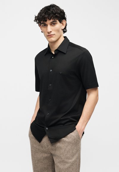 Νεαρός άνδρας με σγουρά μαλλιά, φορώντας μαύρο πουκάμισο με κοντά μανίκια και κολλητά καφέ παντελόνια, με τα χέρια στις τσέπες, ουδέτερο φόντο.