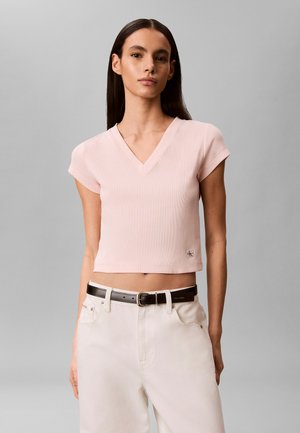 Femme aux longs cheveux foncés porte un crop top rose clair à manches courtes en V et un pantalon blanc avec ceinture noire sur fond gris.
