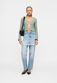 Γυναίκα με πλεγμένα μαλλιά, που φοράει πράσινη κοντή ζακέτα, ανοιχτό μπλε στρωμένη τζιν φούστα και τζιν παντελόνι, μαύρες γόβες και μαύρη τσάντα ώμου.