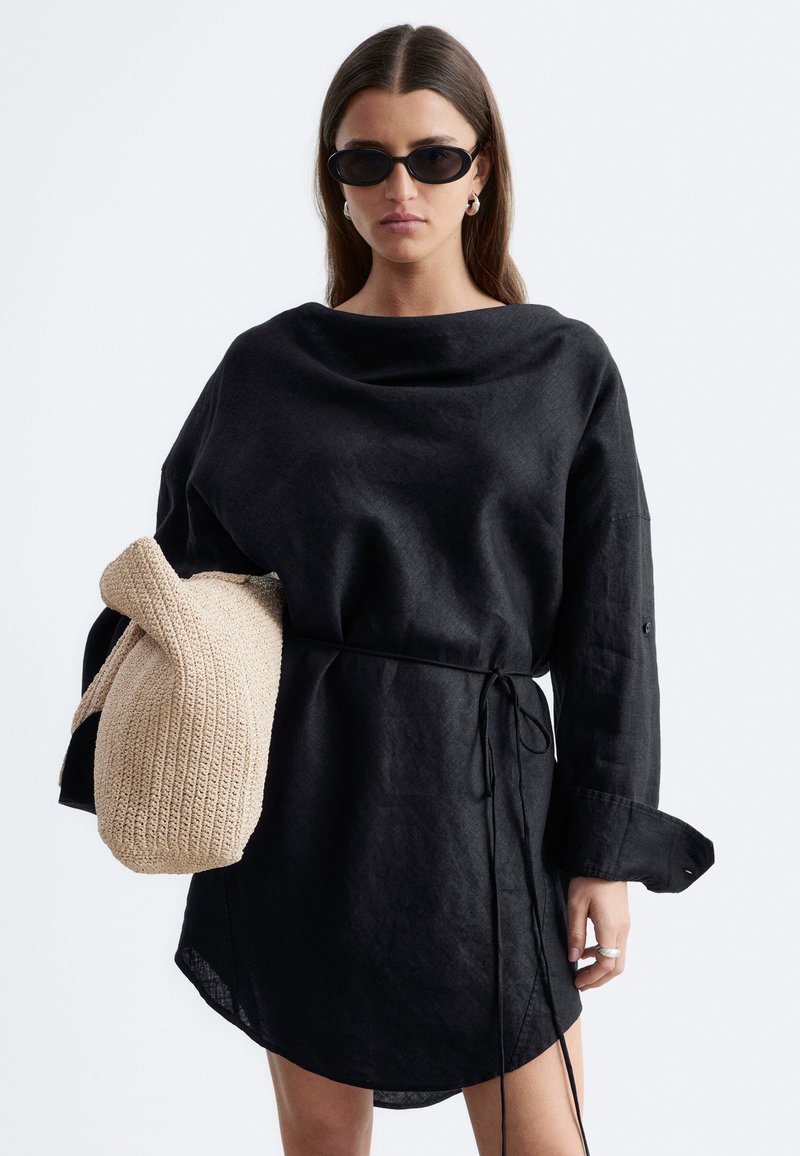 Femme portant une robe noire oversize avec une taille cintrée, des lunettes de soleil noires, tenant un sac beige en tricot sur un fond clair uni.