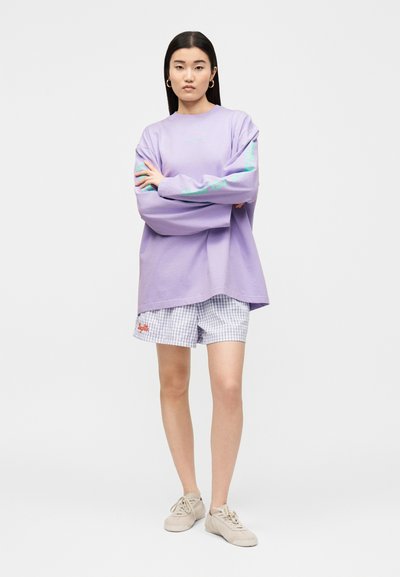 Νεαρή γυναίκα με μακριά μαύρα μαλλιά στέκεται με τα χέρια σταυρωμένα, φορώντας ένα υπερμεγέθες λιλά φούτερ, καρό σορτς και μπεζ αθλητικά παπούτσια.