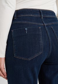 Gros plan sur une poche de jean en denim bleu foncé avec un détail de bouton, porté avec un haut à manches longues blanc sur le côté gauche.