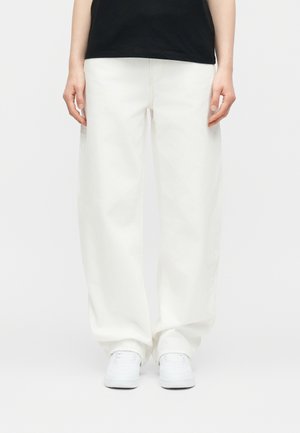 Henkilö pukeutuneena löysiin valkoisiin housuihin, valkoisiin lenkkareihin ja mustaan paitaan, seisoo yksinkertaisen vaalean taustan edessä.