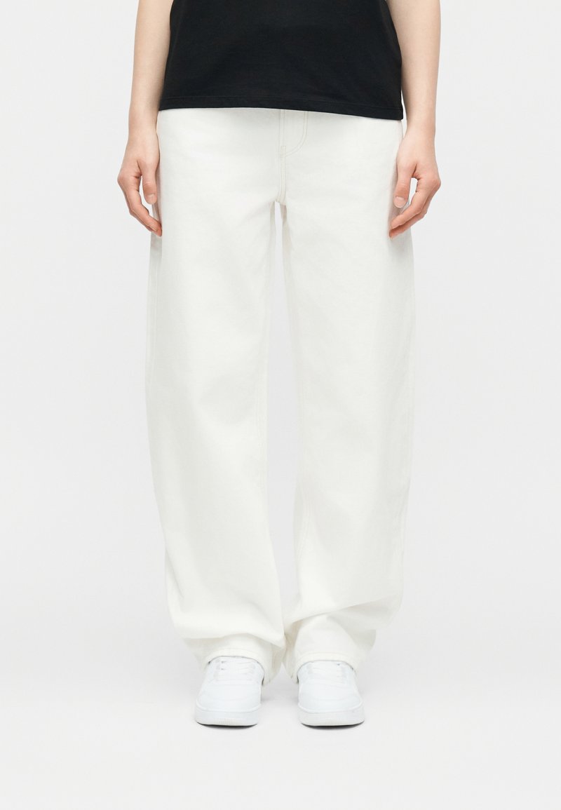 Άτομο που φοράει άνετο λευκό παντελόνι, λευκά αθλητικά παπούτσια και μαύρο μπλουζάκι, στέκεται μπροστά σε απλό φωτεινό φόντο.