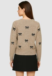 Maglione lavorato a maglia beige con motivi a fiocco neri, colletto a girocollo e orlo a costine, abbinato a una gonna nera. La texture sembra morbida.