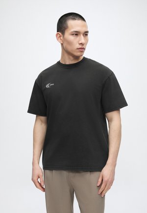 Άνδρας με κοντά μαλλιά που φοράει μαύρο t-shirt με λογότυπο ARKK και μπεζ παντελόνι, κοιτάζει προς τα αριστερά σε απλό φωτεινό φόντο.