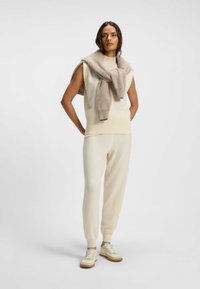 Beige sleeveless knit top with ribbed hem, paired with cream tapered pants and white sneakers. A light brown scarf drapes over the shoulders.