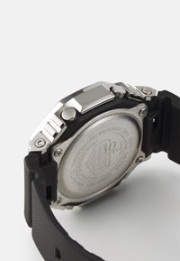 Montre numérique avec bracelet en caoutchouc noir, boîtier en acier inoxydable et fond en argent. Présente des gravures et des boutons sur le côté pour la fonctionnalité.