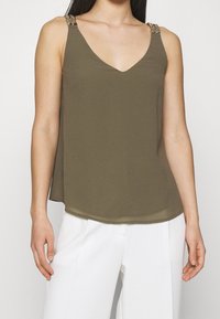 Top sin mangas de color verde oliva con escote en V y tirantes decorativos. Textura suave y fluida con un dobladillo curvado. Combinado con pantalones blancos.