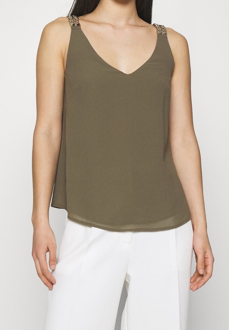 Top sin mangas de color verde oliva con escote en V y tirantes decorativos. Textura suave y fluida con un dobladillo curvado. Combinado con pantalones blancos.