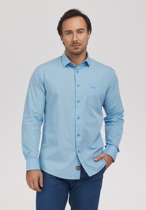 Mężczyzna w jasnoniebieskiej, długorękawowej koszuli zapinanej na guziki i ciemnoniebieskich spodniach, stojący z jedną ręką w kieszeni na jednolitym tle.