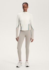 Jachetă albă cu fermoar și accente turcoaz, asociată cu colanți bej și pantofi sport albi. Textură netedă; croială și design athletic.