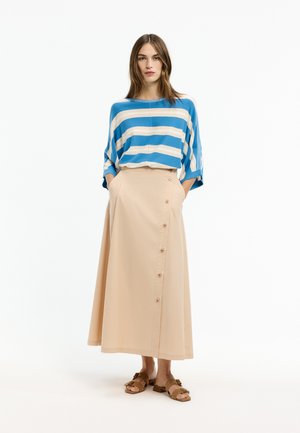 Frau trägt ein blau-weiß gestreiftes lockeres Oberteil, einen beige farbenen, knöpfbaren Midi-Rock mit Taschen und braune Slipper-Sandalen und steht vor weißem Hintergrund.