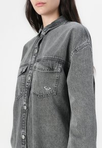 Grå denimskjorte med knappelukking foran, spiss krage og to brystlommer. Har logo-brodere og en avslappet passform.