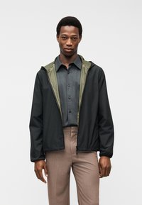 Άνδρας που φοράει μαύρο μπουφάν με φερμουάρ και επένδυση ελιάς πάνω από γκρι πουκάμισο με κουμπιά και καφέ παντελόνι, στέκεται μπροστά σε λευκό φόντο.