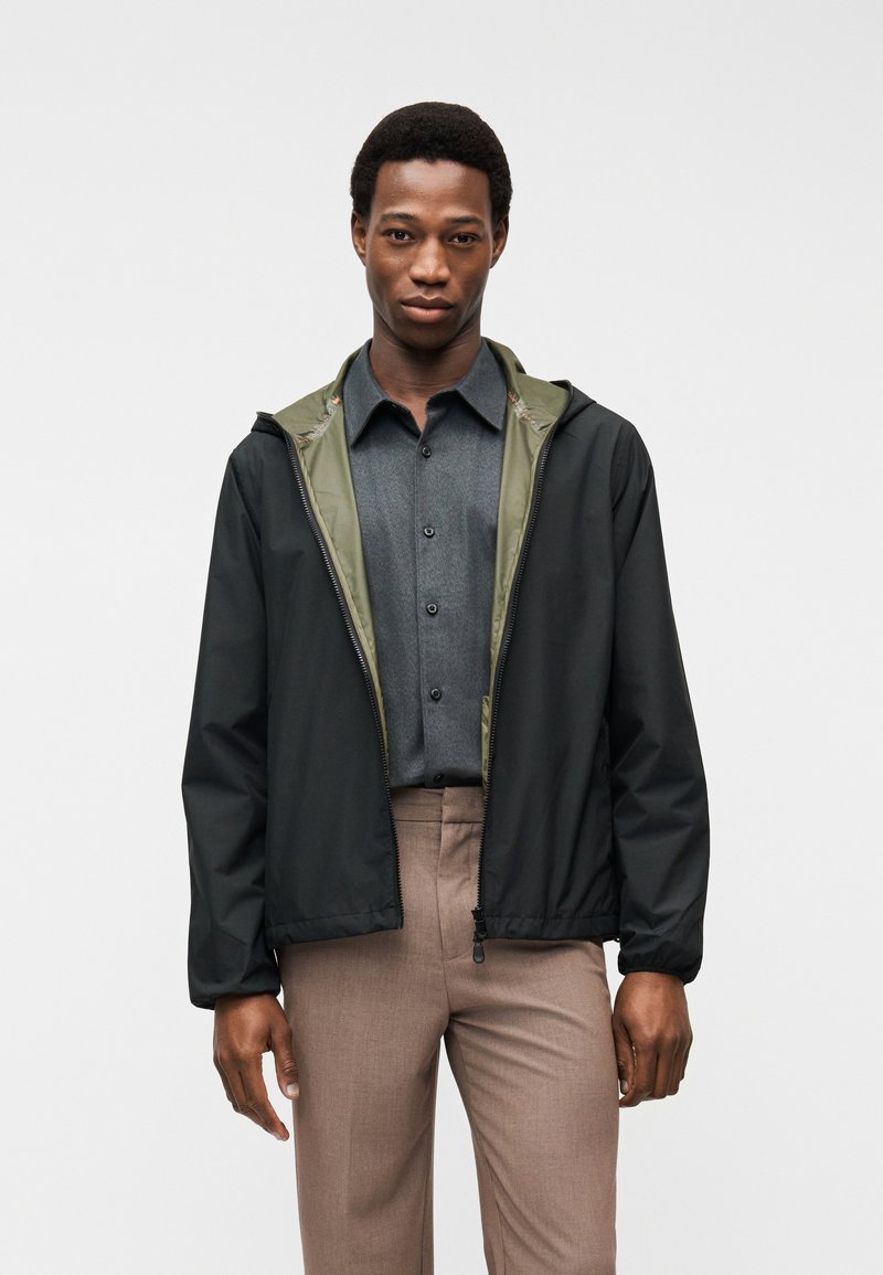 Άνδρας που φοράει μαύρο μπουφάν με φερμουάρ και επένδυση ελιάς πάνω από γκρι πουκάμισο με κουμπιά και καφέ παντελόνι, στέκεται μπροστά σε λευκό φόντο.