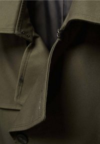 Olivgrüne Jacke mit glatter Textur, ausgestattet mit schwarzen Knöpfen, einem Reißverschluss und einem sauber genähten Kragen. Das Innere zeigt ein dunkles Futter.