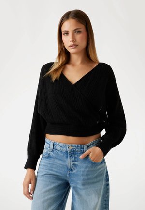 Young woman with straight light brown hair wears a black V-neck cropped sweater and light blue high-waisted jeans, one hand in pocket.