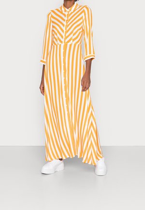 Person trägt ein langes, gelb-weiß gestreiftes Hemdblusenkleid mit dreiviertel Ärmeln und weiße Plateausneaker.