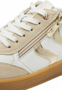 Baskets en daim beige, avec des panneaux en cuir blanc, des accents dorés, une fermeture éclair et des lacets blancs. La semelle est texturée et de couleur gomme.