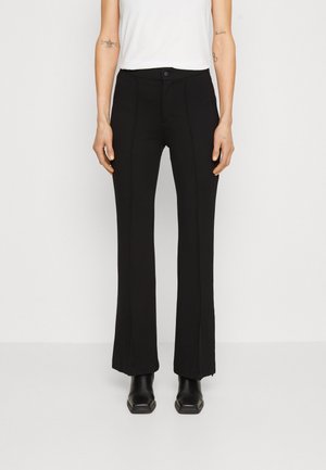Femme portant un pantalon noir droit, des bottes noires et un haut blanc sans manches sur un fond uni.