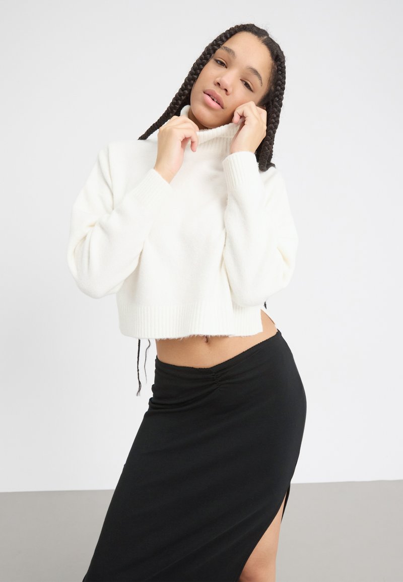 Weißer, kurz geschnittener Pullover mit hohem Kragen und gerippten Bündchen, kombiniert mit einem schwarzen Rock mit seitlichem Schlitz. Weicher Stoff, lässiges Design.