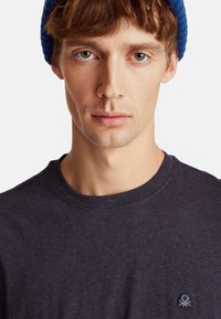 Mladý muž s světlou pletí a hnědými vlasy, oblečený v modré pletené čepici a tmavém tričku s kruhovým výstřihem, na kterém je malá vyšitá nášivka na hrudi.