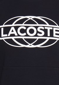Texte blanc "LACOSTE" à l'intérieur d'anneaux elliptiques qui se croisent sur un fond en tissu noir, probablement un sweat-shirt ou un hoodie.
