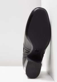 Talons et semelle d'une chaussure habillée en cuir noir visibles, inclinés contre un mur blanc et un sol blanc.