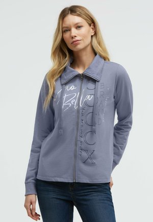 Woman with long blonde hair wearing a blue zip-up jacket with graphic text and dark jeans, standing with one hand in pocket.