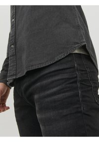 Camisa de mezclilla negra con dobladillo ligeramente curvado, con botones de presión metálicos y una textura sutil, combinada con jeans oscuros.