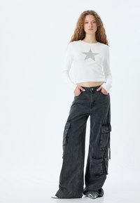 Hvid langærmet crop top med et sølvstjernedesign; parret med brede sorte cargo-bukser, der har flere lommer og en afslappet pasform.