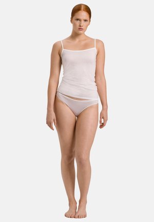 Jeune femme aux cheveux roux portant un caraco blanc et des sous-vêtements, debout pieds nus devant un fond clair uni.