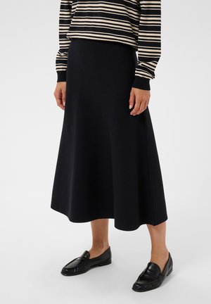 Personne portant une jupe midi noire, des mocassins noirs et un haut à manches longues rayé noir et beige, debout sur un fond blanc.