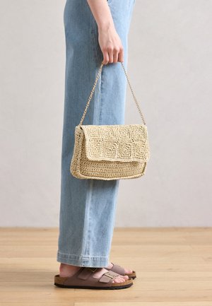 Woven straw handbag with a flap, featuring a geometric pattern, gold chain strap, and a light beige color. Paired with light denim jeans.