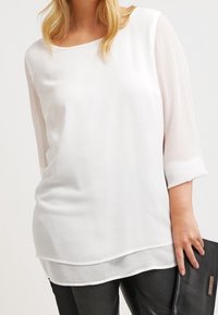 Top blanco con escote redondo y mangas transparentes de tres cuartos, diseño en capas, textura suave, combinado con pantalones de imitación cuero negros.