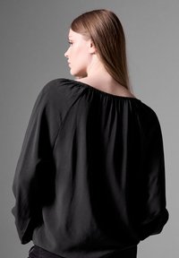 Loose-fitting black blouse with gathered neckline, long sleeves, and soft, textured fabric. The design is simple and elegant, with a subtle sheen.
