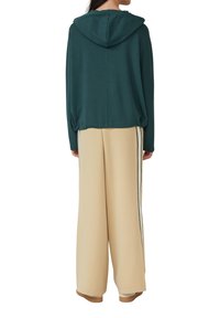 Persona vestita con una felpa con cappuccio verde scuro e pantaloni beige a gamba larga con strisce nere ai lati, in piedi di spalle su uno sfondo bianco.