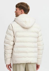 Puffjakke i kremfarge, med hette, quiltet tekstur og elastiske mansjetter. Enkelt design uten synlige logoer eller mønstre.