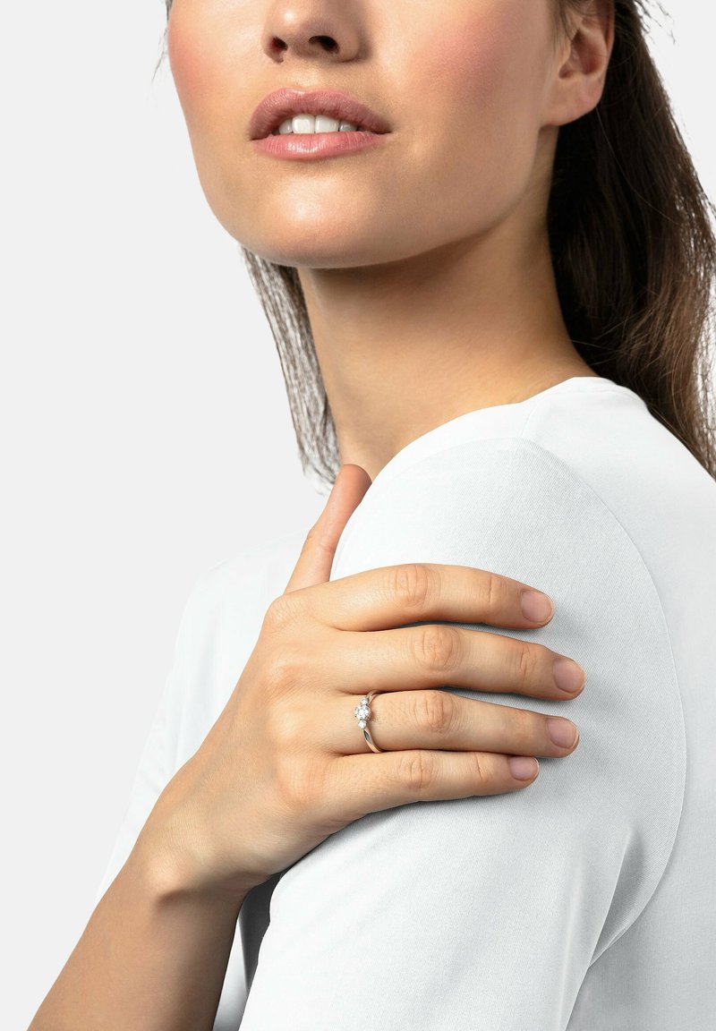 Silberner Ring mit drei klaren Edelsteinen, der an einer Hand zu sehen ist, die eine Schulter berührt, während ein weißes, eng anliegendes Hemd getragen wird. Glatte Textur, einfaches Design.