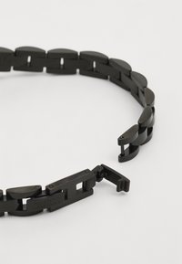 Juodas metalinis apyrankė, turinti ovalius žiedus su lygiu paviršiumi, išskirtinį užsegimą ir „GUESS“ logotipą išgraviruotą ant žiedo.
