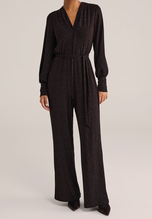 Vrouw draagt donkerbruine lange mouwen jumpsuit met V-hals, taille met riem, wijde pijpen en zwarte puntige schoenen, staand op een neutrale achtergrond.