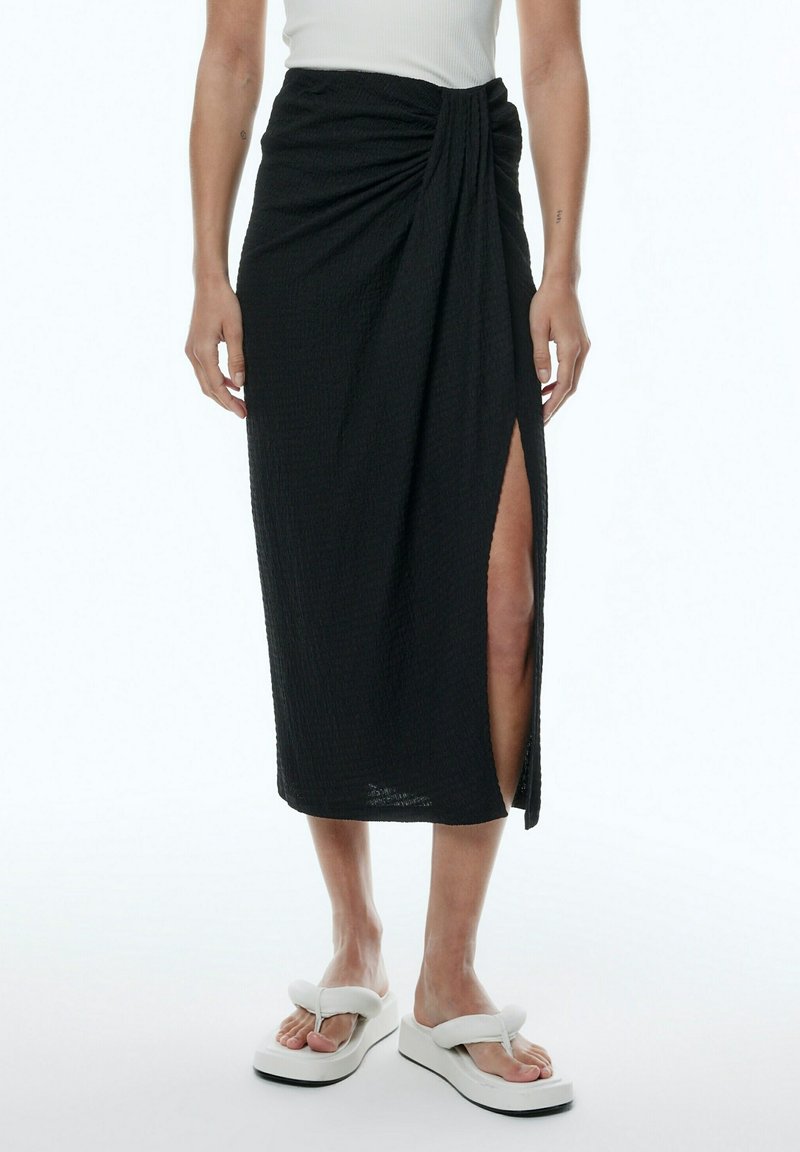 Femme portant une jupe midi noire avec un nœud à l'avant et une fente haute sur la cuisse, associée à des tongs à plateforme blanches rembourrées.